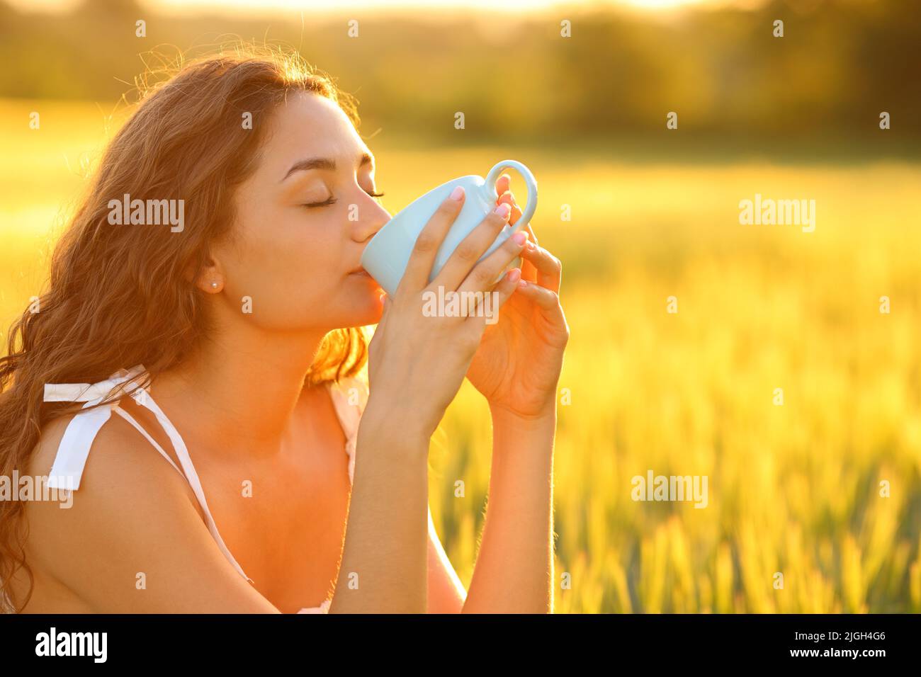 Donna che si rilassa bevendo caffè in un campo di grano al tramonto con una luce calda Foto Stock