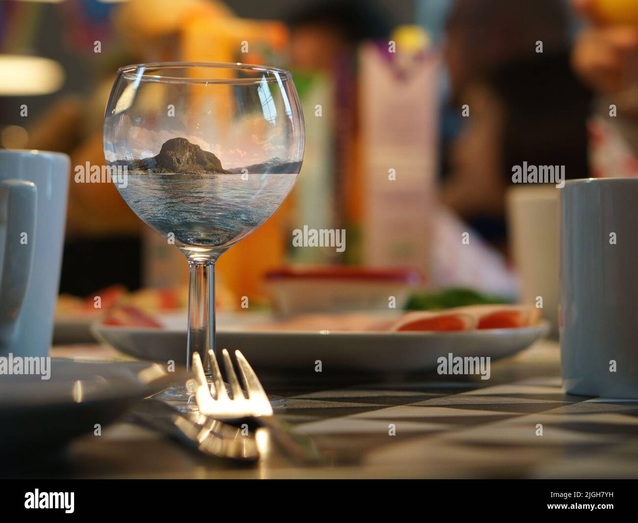 Immagine fantasy che compone un bicchiere di vino con il paesaggio dalla Norvegia vicino al mare nel bicchiere. Su un tavolo da pranzo con posate, piatti e tazze. Creat Foto Stock