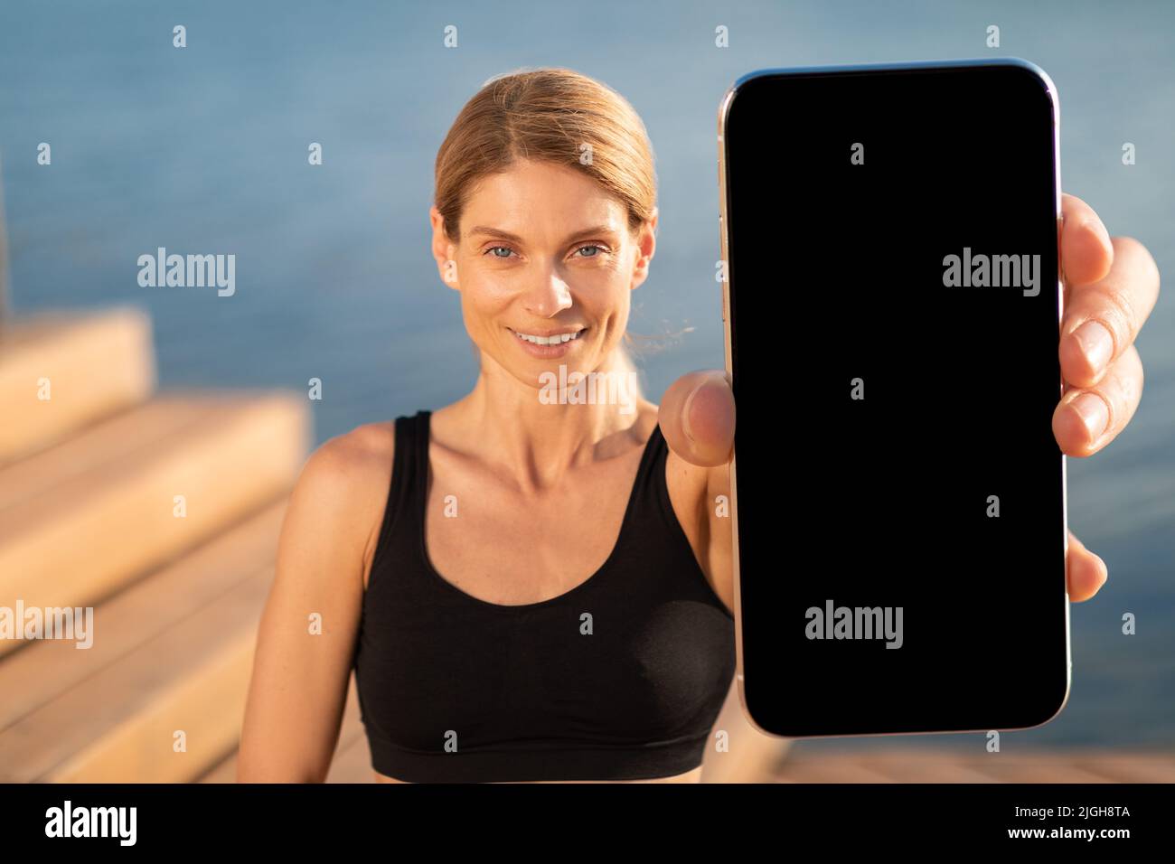 App per il fitness. Donna sportiva di mezza età che mostra grande smartphone vuoto alla fotocamera Foto Stock