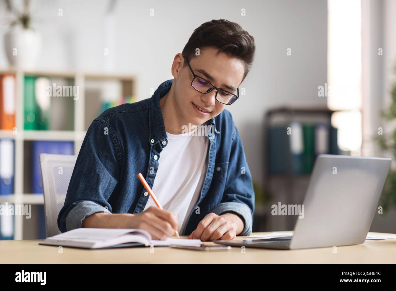 Giovane dipendente di sesso maschile che prende appunti mentre lavora con il portatile in ufficio Foto Stock
