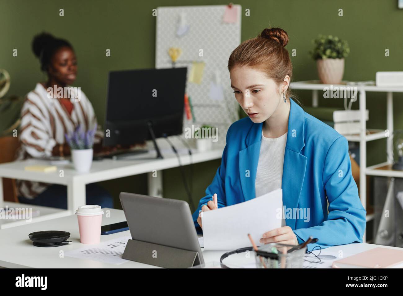 Giovane donna d'affari che guarda lo schermo del tablet pc mentre prende appunti nel documento durante la conversazione online sul suo posto di lavoro Foto Stock