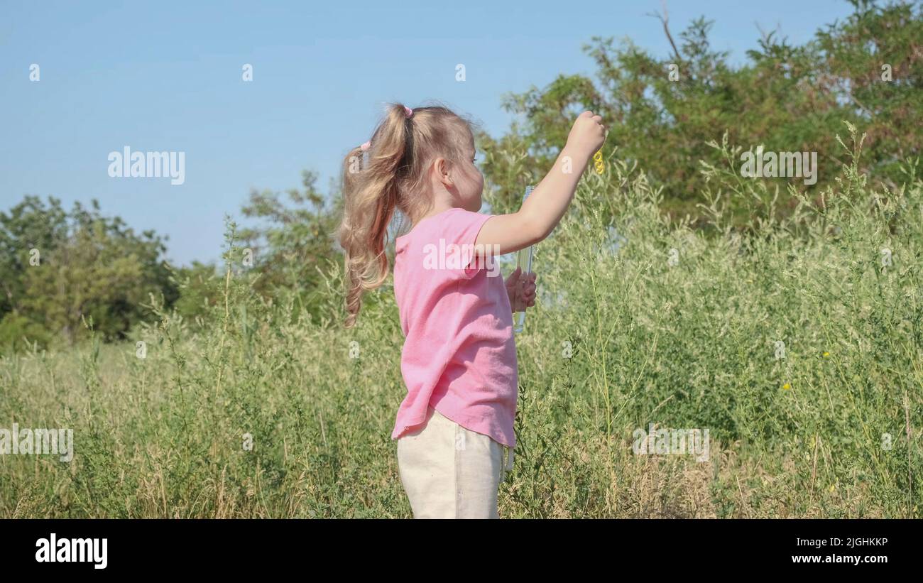 Primo piano ritratto di bambina gioca con bolle di sapone in erba alta nel parco. Carino ragazza piccola e soffia le bolle di sapone nel prato il giorno del sole. Foto Stock