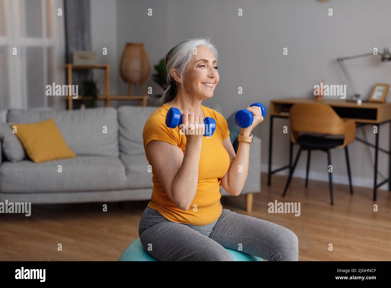 Donna anziana felice esercitandosi con i dumbbells nel paese, seduta sul fit ball e rinforzando i suoi muscoli del braccio Foto Stock