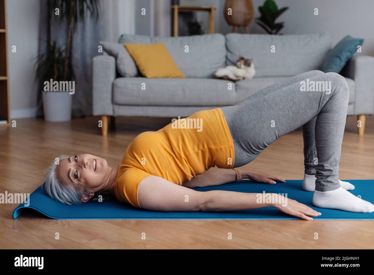 Donna sportiva anziana che fa posa yoga mezzo ponte, rinforzando i suoi muscoli abs, formazione in salotto, spazio copia Foto Stock