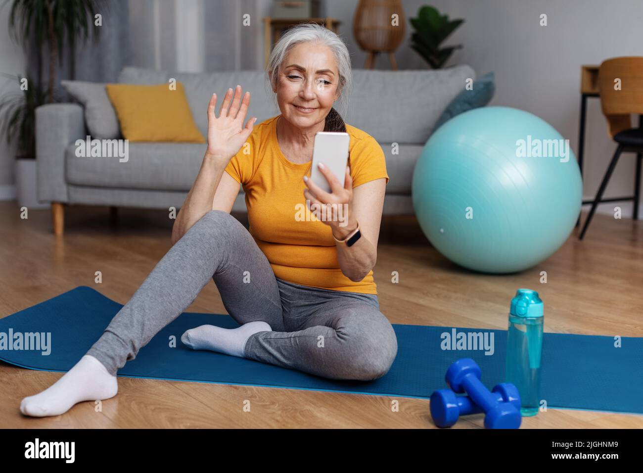 Donna anziana con smartphone che effettua videochiamate con il proprio divano mentre si siede sul materassino fitness a casa Foto Stock