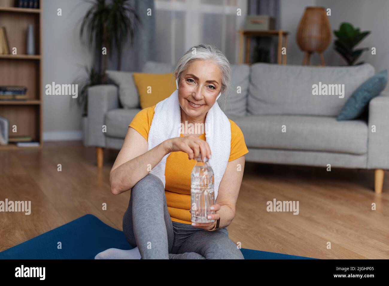 Donna anziana in abbigliamento sportivo seduto su tappeto sportivo con bottiglia d'acqua limpida, che mantiene idratato durante l'allenamento domestico Foto Stock