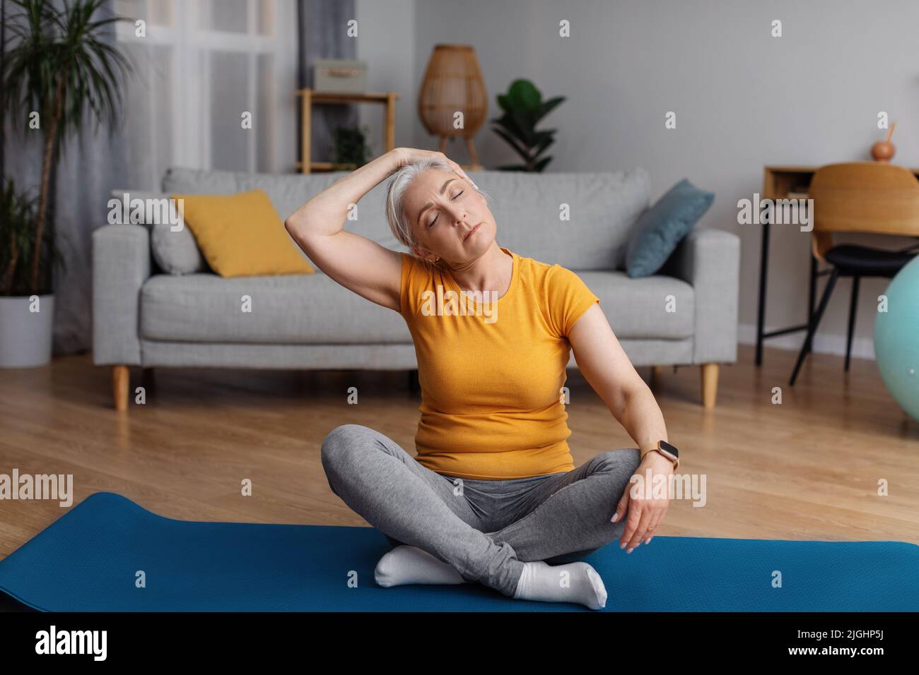 Concetto di stile di vita sano. Donna anziana stretching collo, seduta su tappeto yoga e riscaldamento prima di allenamento domestico Foto Stock