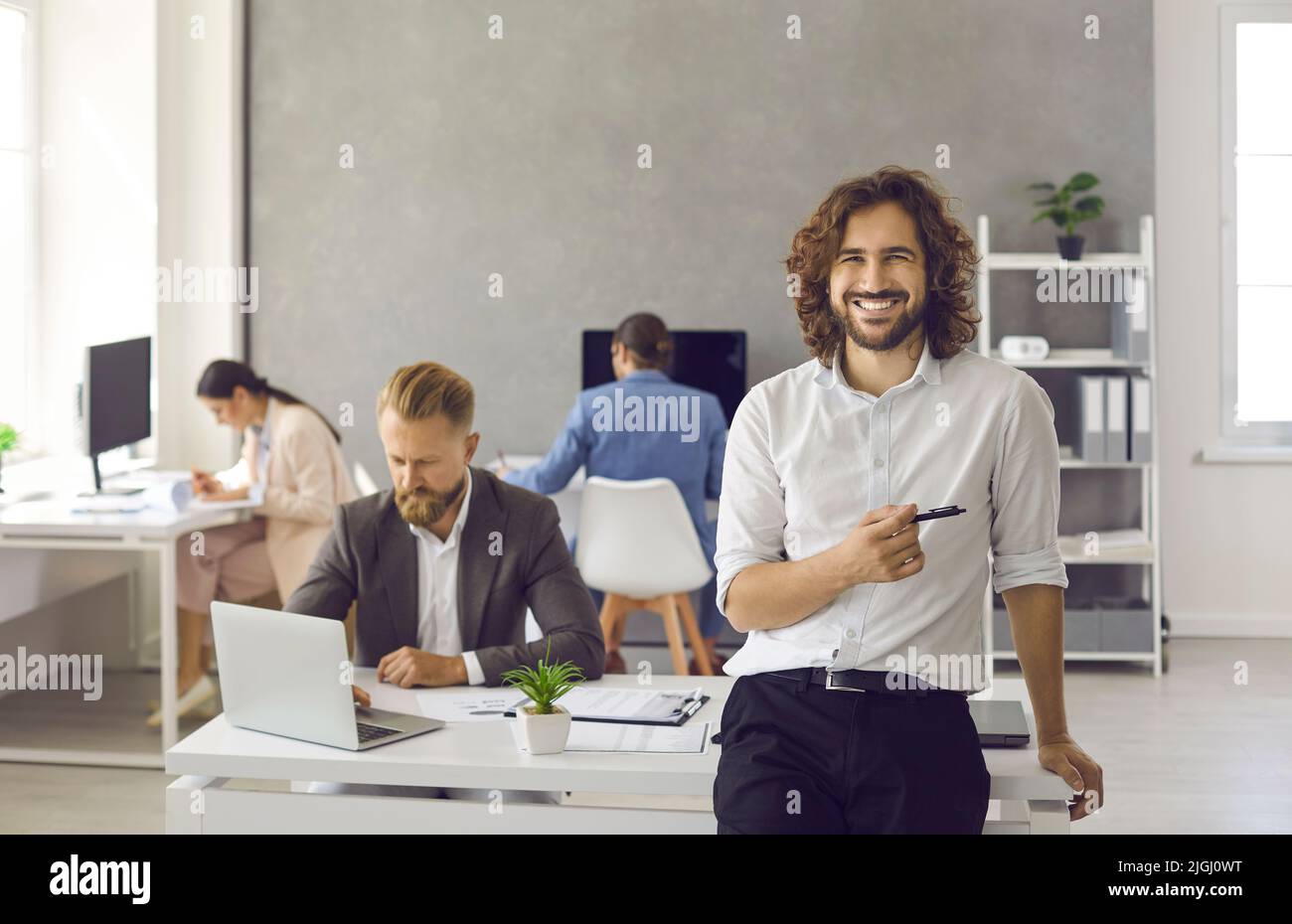 Ritratto del giovane uomo d'affari felice in piedi in ufficio, tenendo la penna e sorridendo Foto Stock