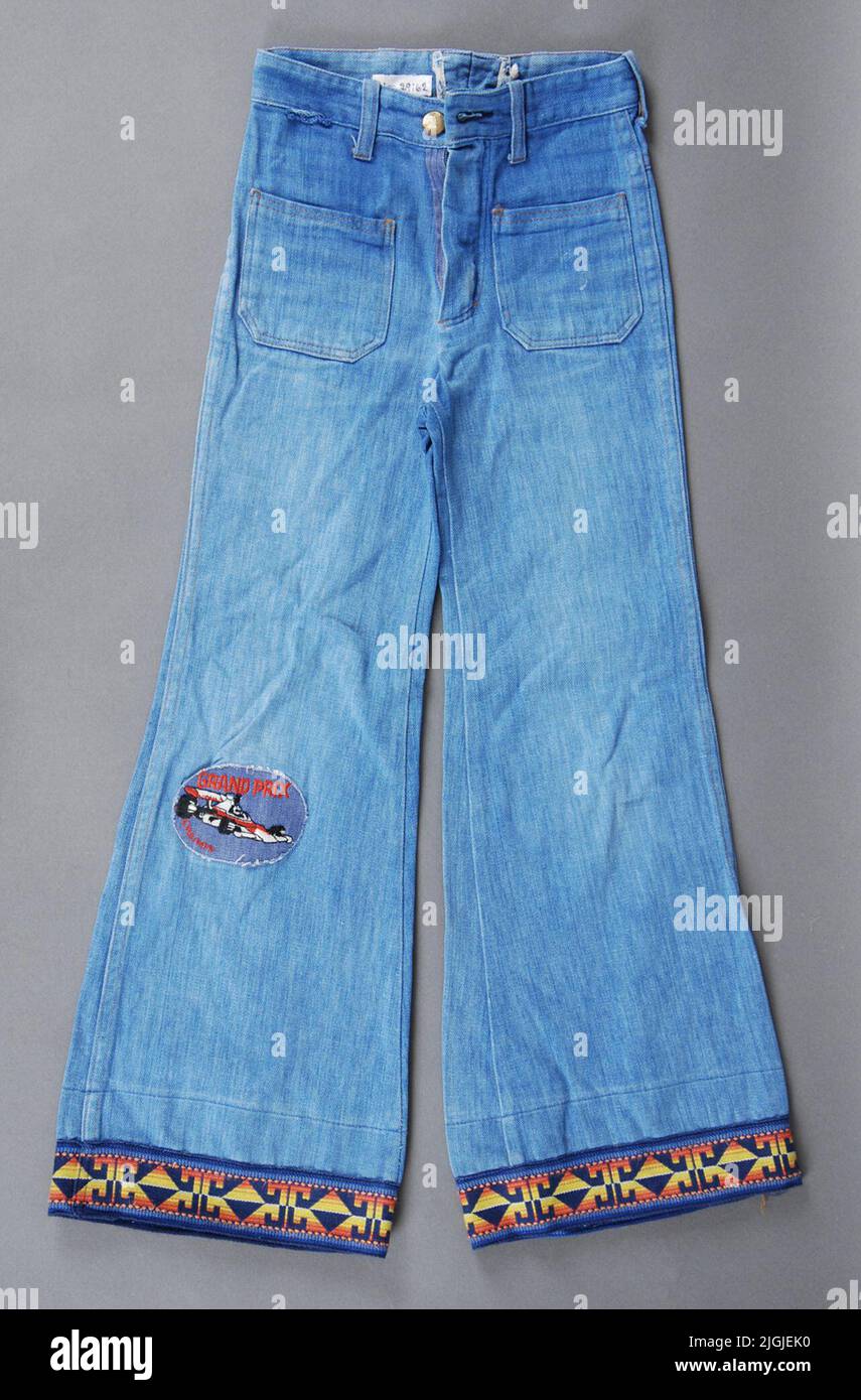 Jeans pantaloni Boy. Jeans in denim, cotone. Pantaloni in vita con gambe svasate. Oro con cerniera. Fodere con bottoni e bottoni in metallo giallo al centro. 6 pezzi per cinghia. Tasche quadrate cucite. Cuciture decorative gialle. Fasce decorate con interruttore sulla parte inferiore delle gambe. Motivo geometrico del nastro tessuto modellato in giallo/arancione contro il fondo blu. Sulla gamba destra incollata la squadra con il testo rosso "Grand Prix" e la Formula 1 in rosso, bianco, nero. Dimensioni: C 125. (Marchiatura della vita interna) larghezza della vita 270 mm, Benvidd 230 mm. Appello di Morgan Olsson, Swan Hall 2014 di Morgan Olsson, Svanha Foto Stock