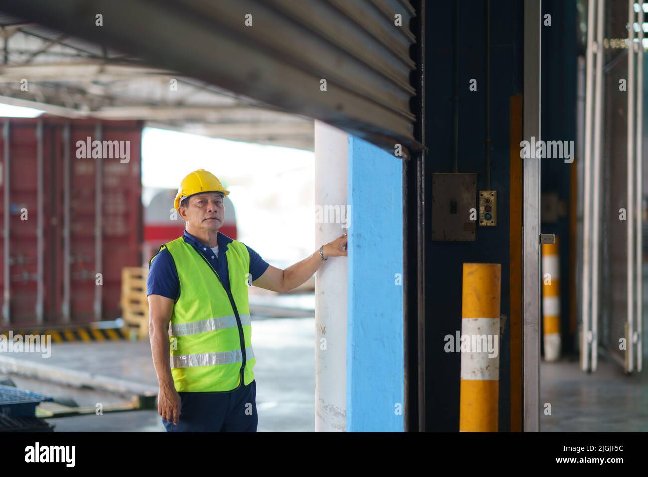 Operatore del magazzino asiatico in tute e casco protettivo che apre la porta a rullo della serranda all'inizio della giornata lavorativa Foto Stock