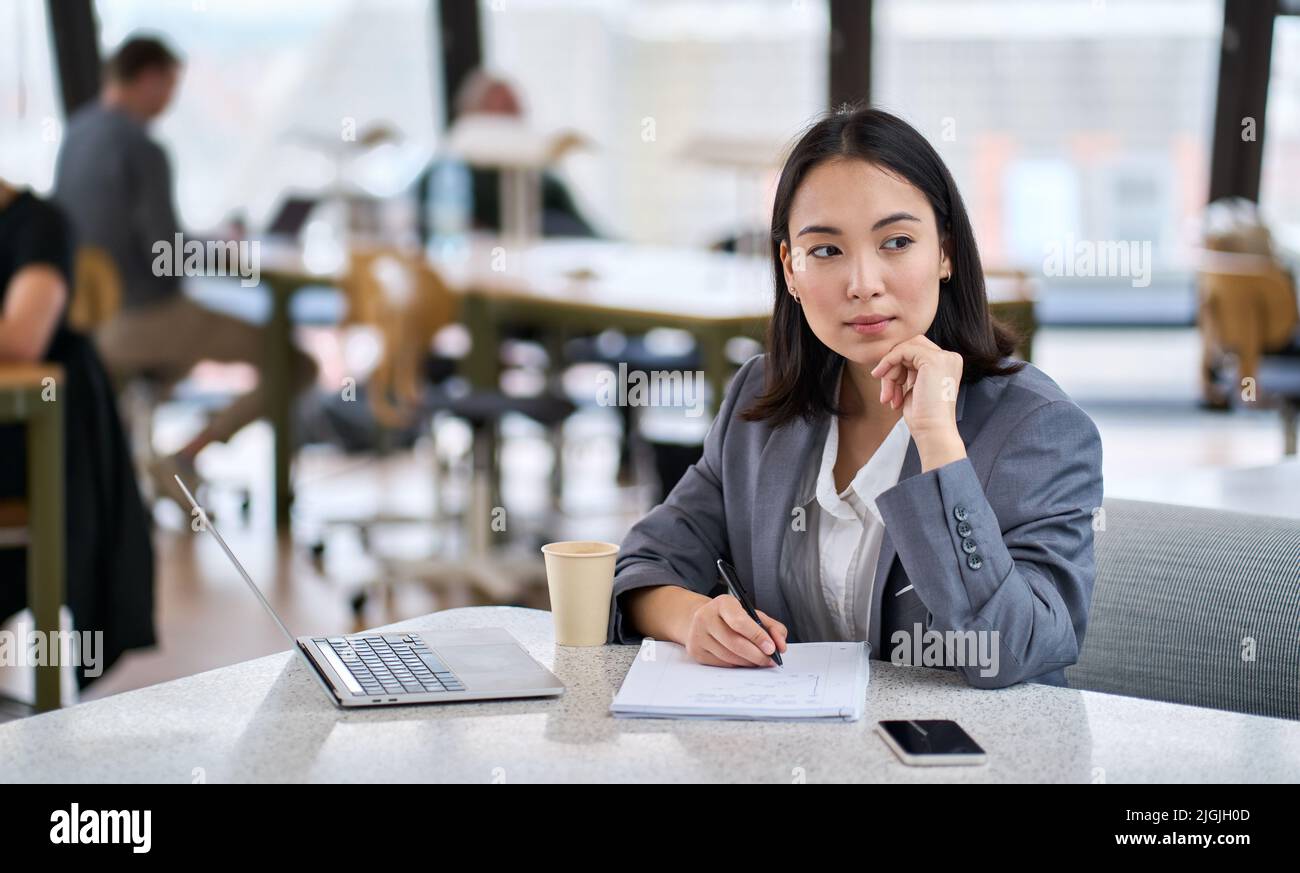 Giovane imprenditrice asiatica che lavora in un ufficio moderno, prendendo appunti e pensando. Foto Stock
