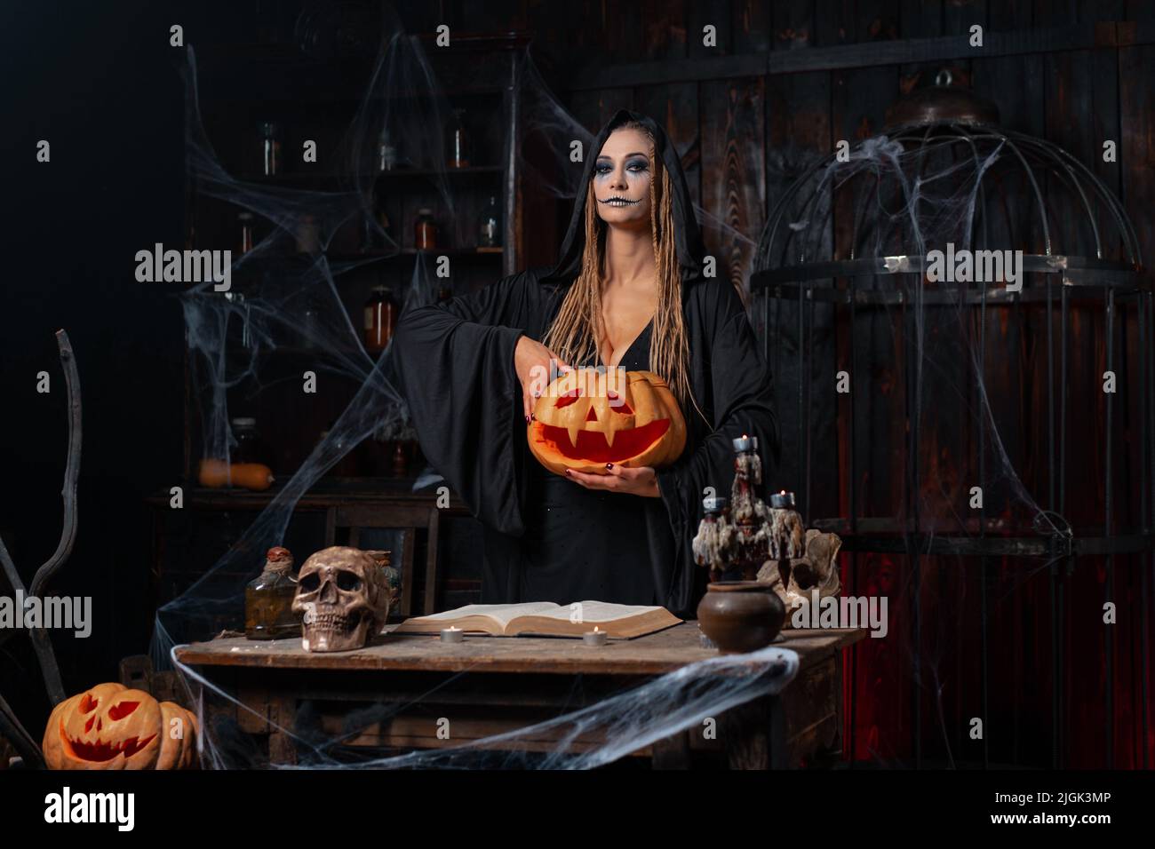 Concetto di Halloween. Strega nera che tiene la zucca di Halloween in mano con il viso smily scolpito in piedi a mano stanza scura. Necromancer femminile, luogo della prigione. Foto Stock