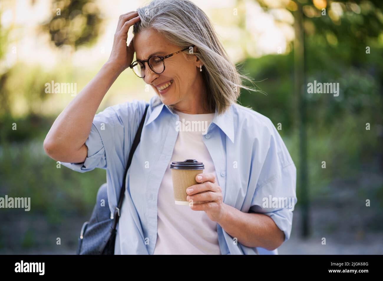 Affascinante donna matura beve caffè in movimento fissando i suoi capelli. Donna matura trascorrere del tempo libero viaggiando nella città vecchia europea gustando una tazza di tè e caffè. Donna dai capelli grigi in casual con zaino. Foto Stock