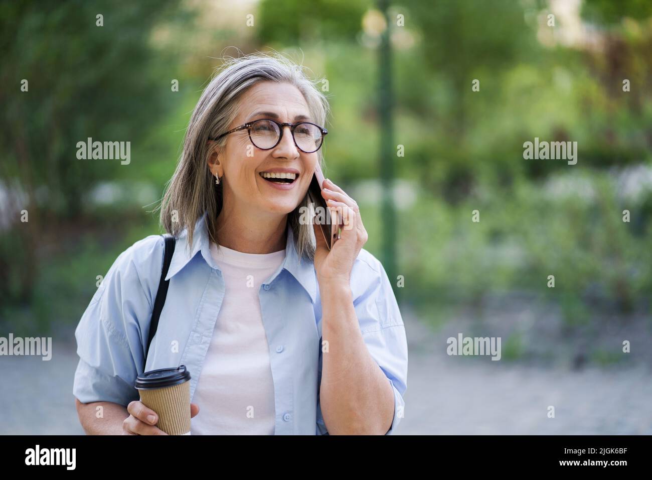 Donna d'affari europea con capelli grigi che parla al telefono mentre beve caffè in movimento utilizzando una tazza di carta nel giardino o nel parco della città. Donna matura alla pausa caffè, con camicia blu e maglietta bianca. Foto Stock
