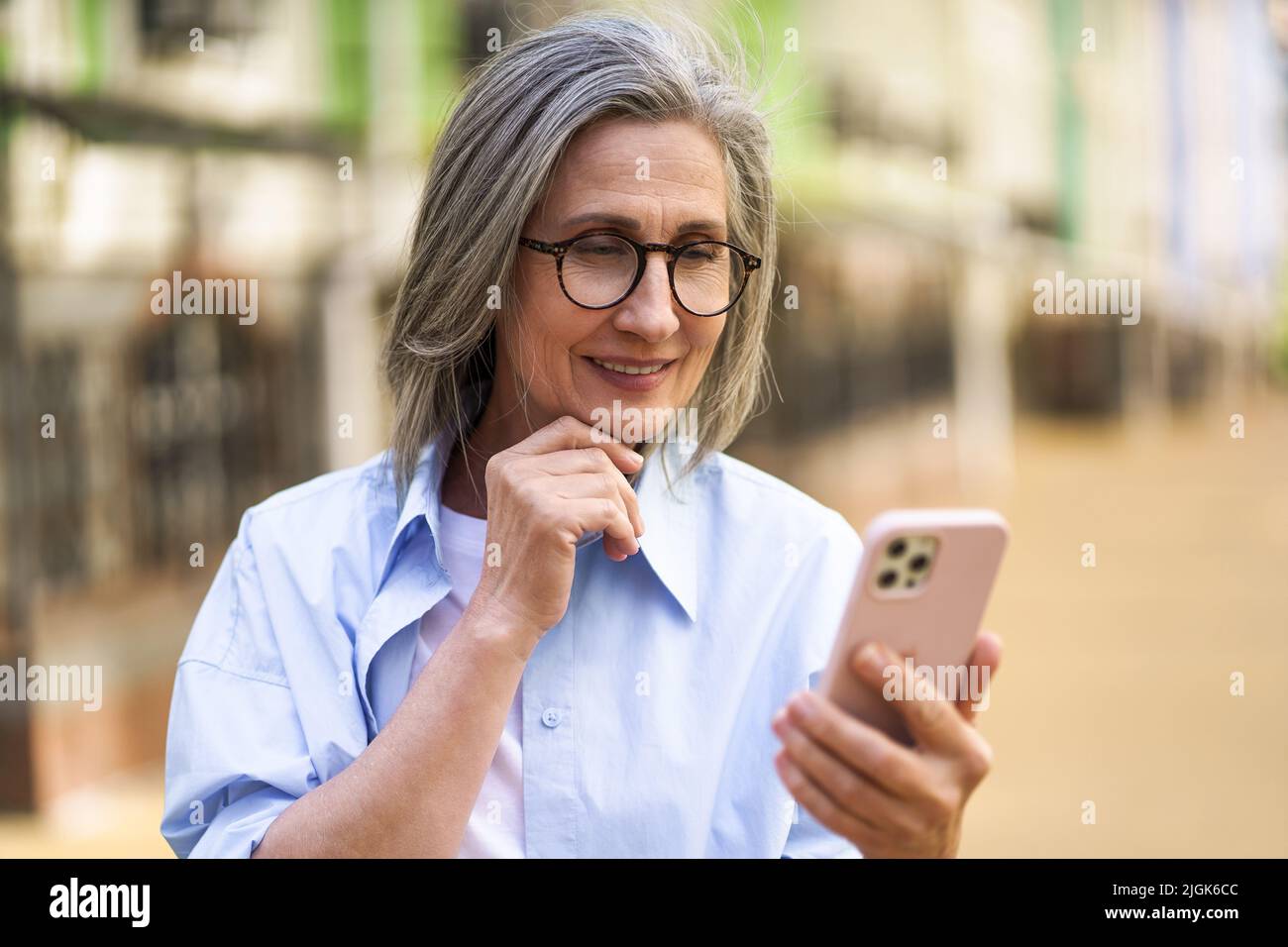 Affascinante donna matura con capelli grigi che tiene smartphone per le strade della città vecchia europea. Una donna matura legge un messaggio di testo o risponde a una videochiamata in piedi o all'aperto. Foto Stock
