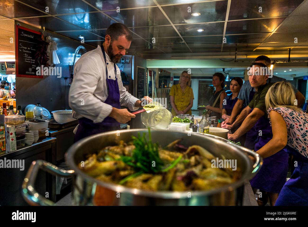 Nel suo corso di cucina nella sala del mercato di Avignon, in Francia, Jon Chiri versa una salsa chiara su una pentola di pollame rosolato mentre i partecipanti guardano. Foto Stock