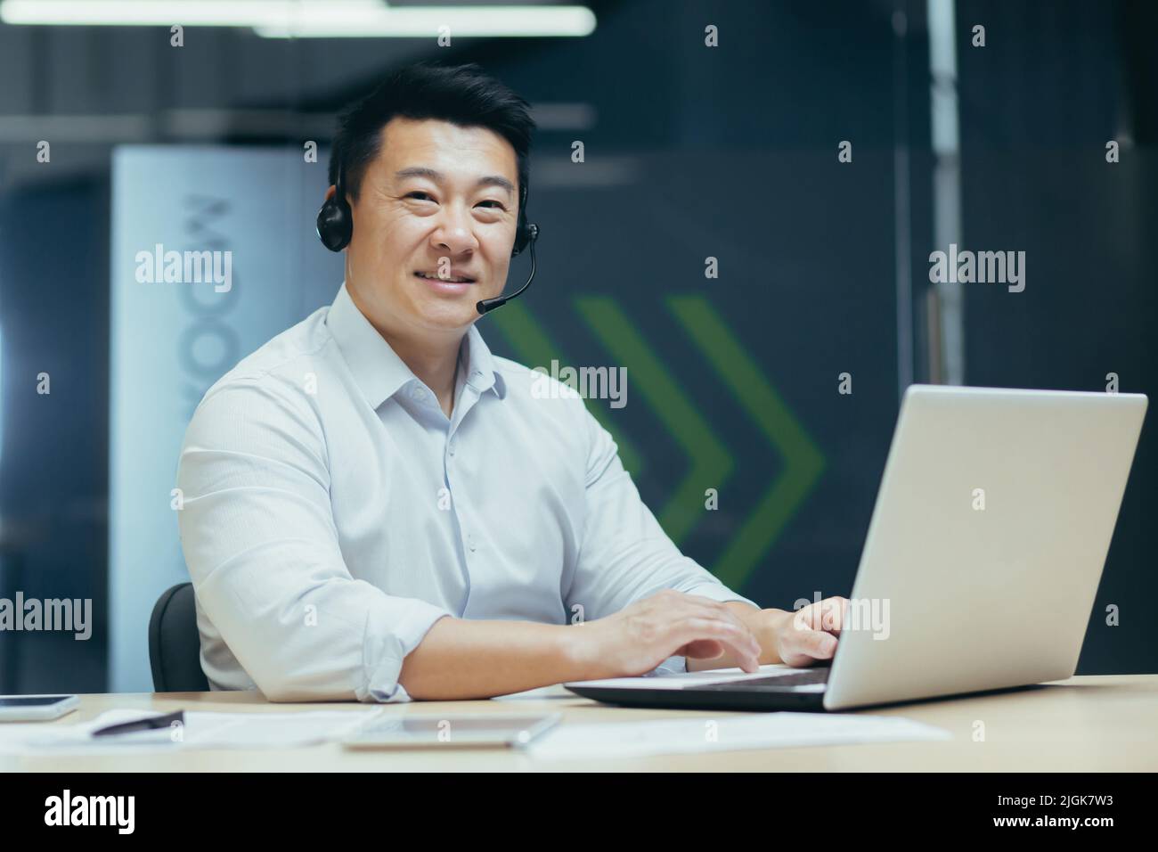 Ritratto di uomo d'affari asiatico di successo con cuffie video chiamata guardando la fotocamera e sorridente, consulente di supporto tecnico che lavora in ufficio con il laptop Foto Stock
