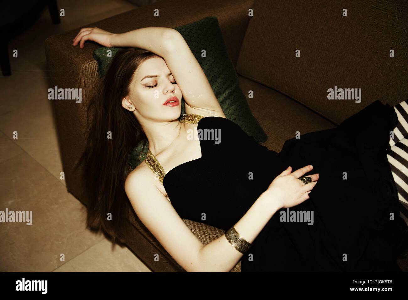 Una bruna che indossa un abito nero corto e leggings a righe è adagiata su un divano, con gli occhi chiusi e un braccio sul bracciolo Foto Stock