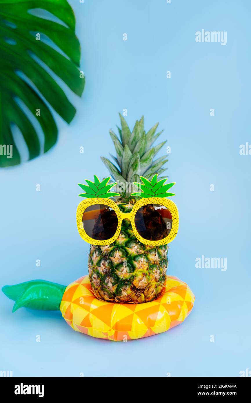 Ananas divertente in un bicchiere di ananas forma sul giallo gonfiabile anello su sfondo blu verticale. Estate in stile tropicale sulle vibrazioni della spiaggia. Foto Stock