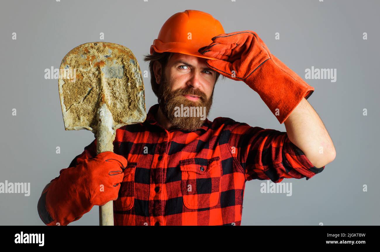 Costruttore sorridente in elmetto con pala. Uomo da lavoro in guanti da lavoro con langa. Costruzione e riparazione. Foto Stock