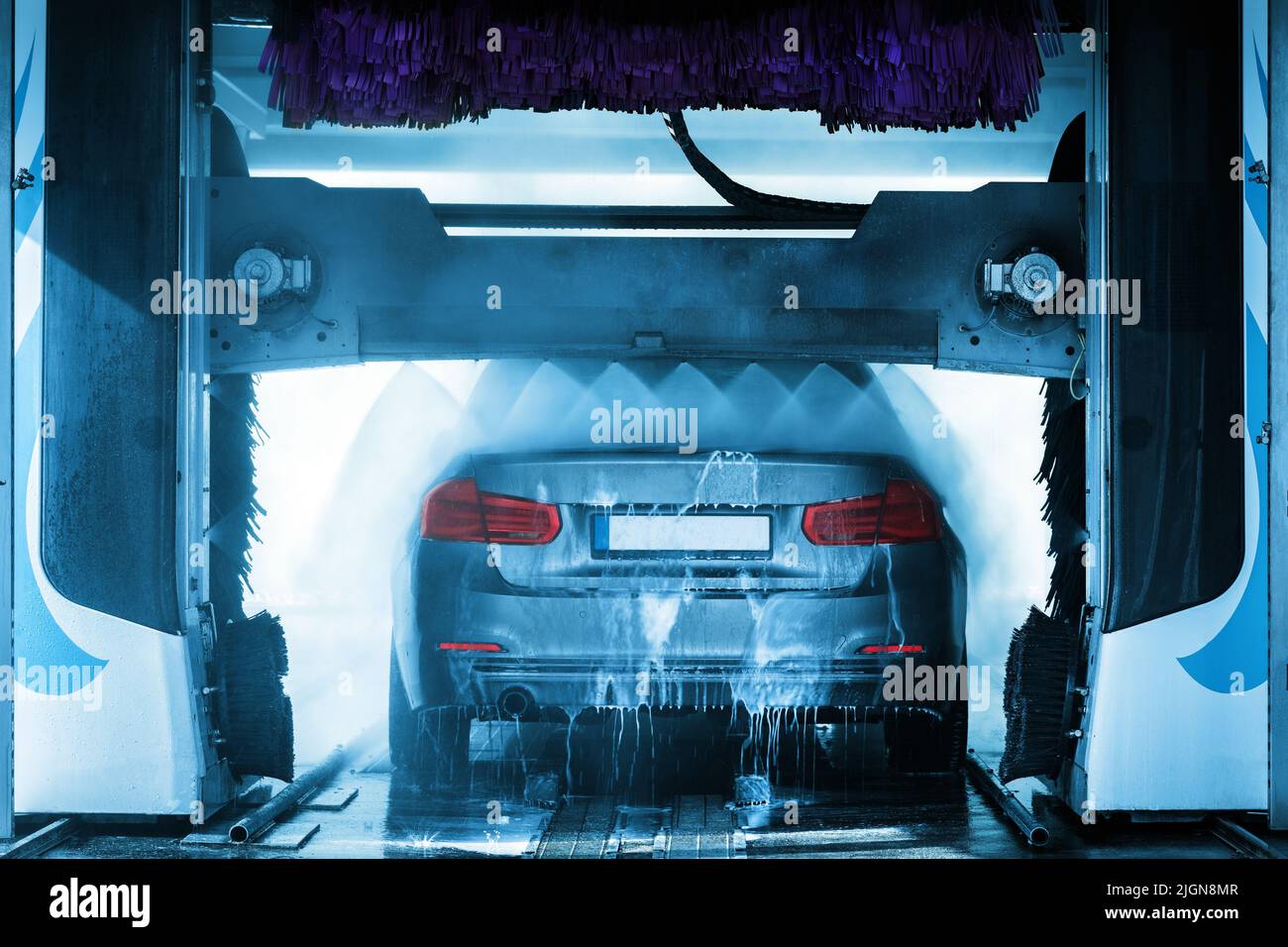 Un'auto grigia che attraversa un autolavaggio automatico con spazzole e acqua che colpiscono il veicolo. Foto Stock