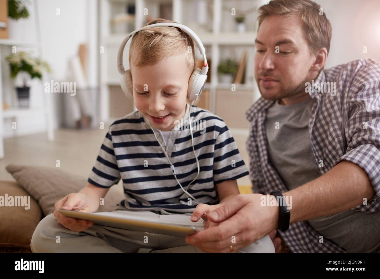 Ragazzo simpatico biondo-haired positivo in cuffie cablate utilizzando l'app di apprendimento sul tablet insieme al padre a casa Foto Stock