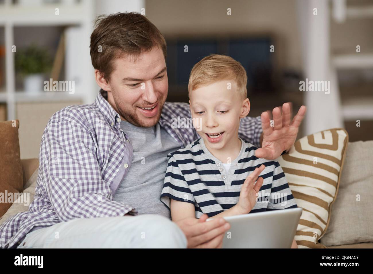 Positivo bel padre e suo figlio seduti sul divano e agitare le mani al tablet mentre parla con la madre tramite l'app di chat video Foto Stock
