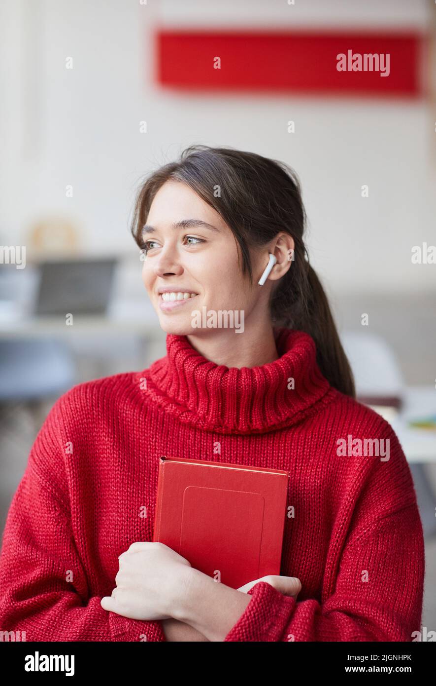 Bella ragazza stilista positiva con auricolari wireless indossando maglia rossa maglione che abbraccia personal planner e guardare via Foto Stock