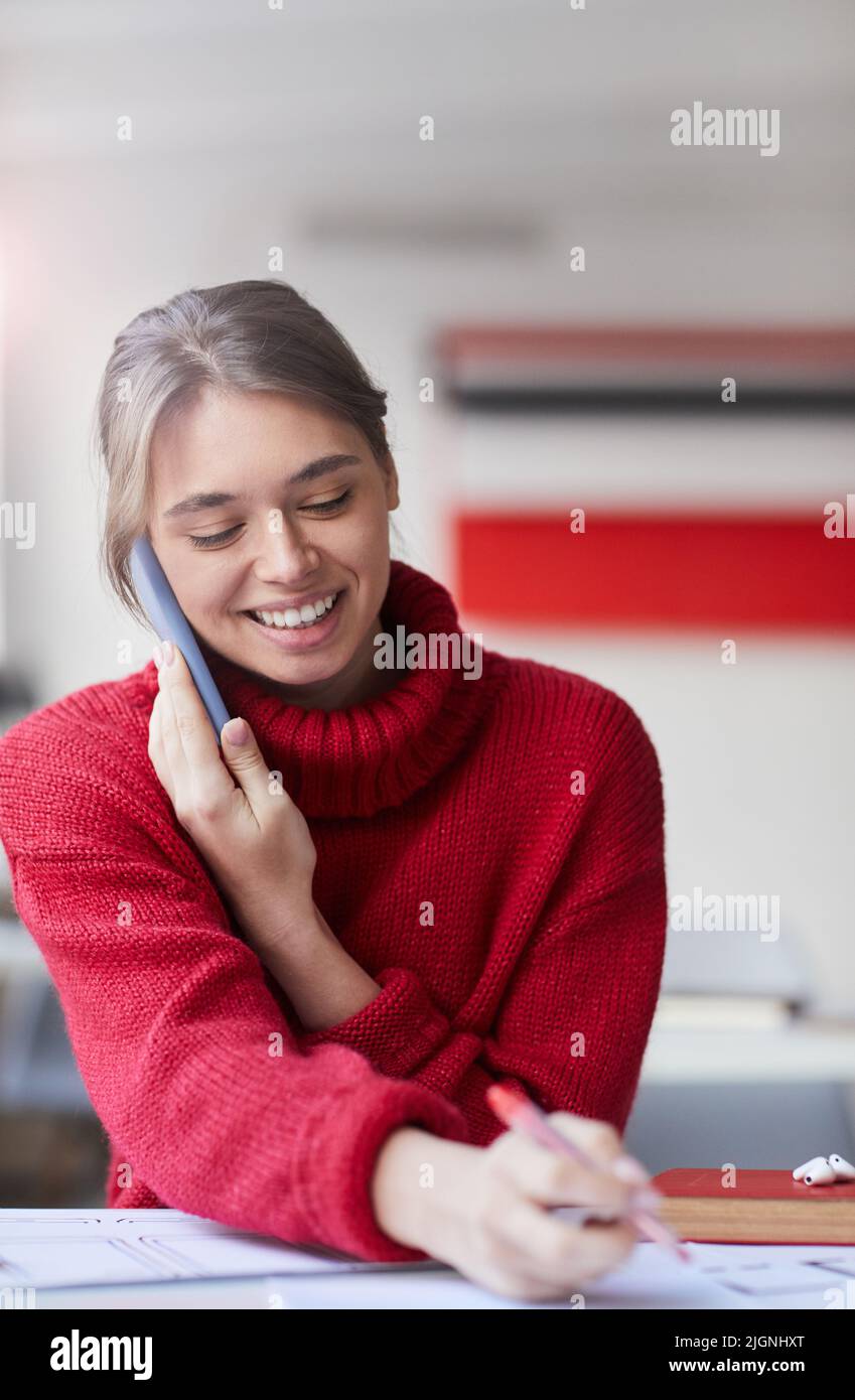 Positivo attraente manager femminile in elegante maglione seduta alla scrivania in ufficio e discutere di UX design con il cliente per telefono Foto Stock