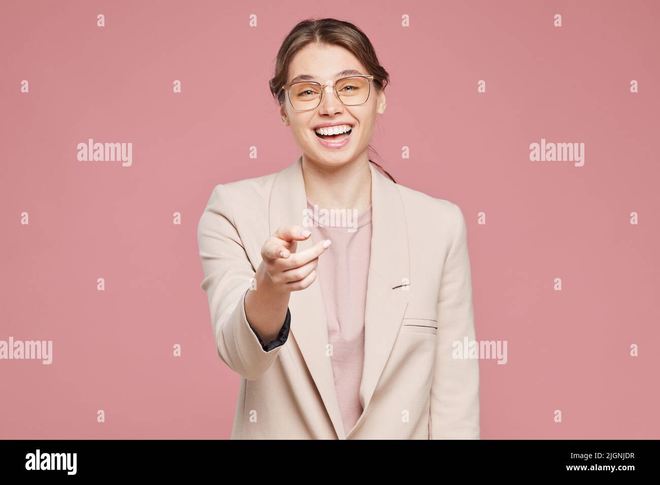 Ritratto di bella ragazza positiva in occhiali eleganti e giacca beige in piedi su sfondo rosa e ridendo mentre punta con il dito contro il camer Foto Stock