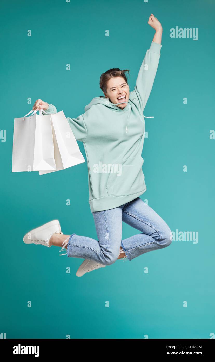 Ritratto di positivo eccitato hipster ragazza in hoodie jumping con acquisti dopo la vendita shopping, teal background Foto Stock