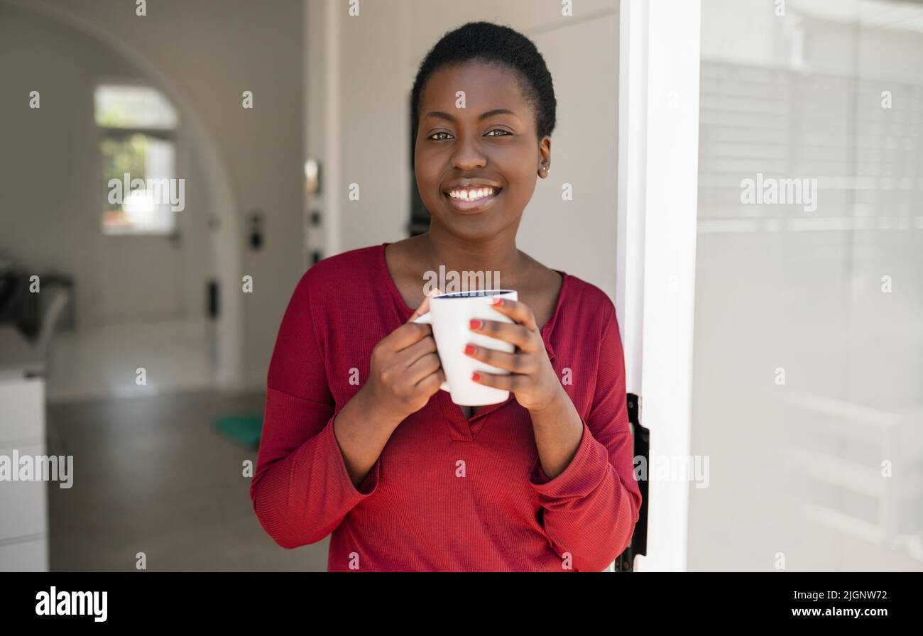 Ritratto di una bella donna africana in piedi in porta modo di casa, tenendo una tazza, sorridendo e guardando in macchina fotografica Foto Stock