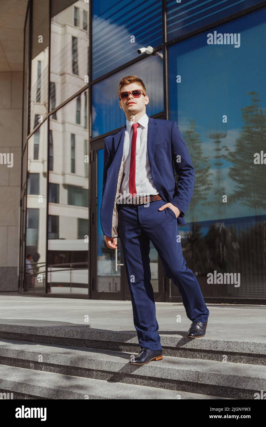 Elegante giovane uomo in abito blu, camicia bianca, cravatta rossa, calzini  blu e scarpe in pelle con occhiali da sole in legno in un moderno edificio  aziendale Foto stock - Alamy