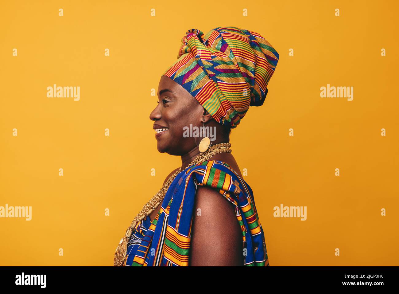Felice donna africana che indossa un abbigliamento tradizionale su sfondo giallo. Donna matura nera vestita in colorato panno Kente e gioielli dorati Foto Stock