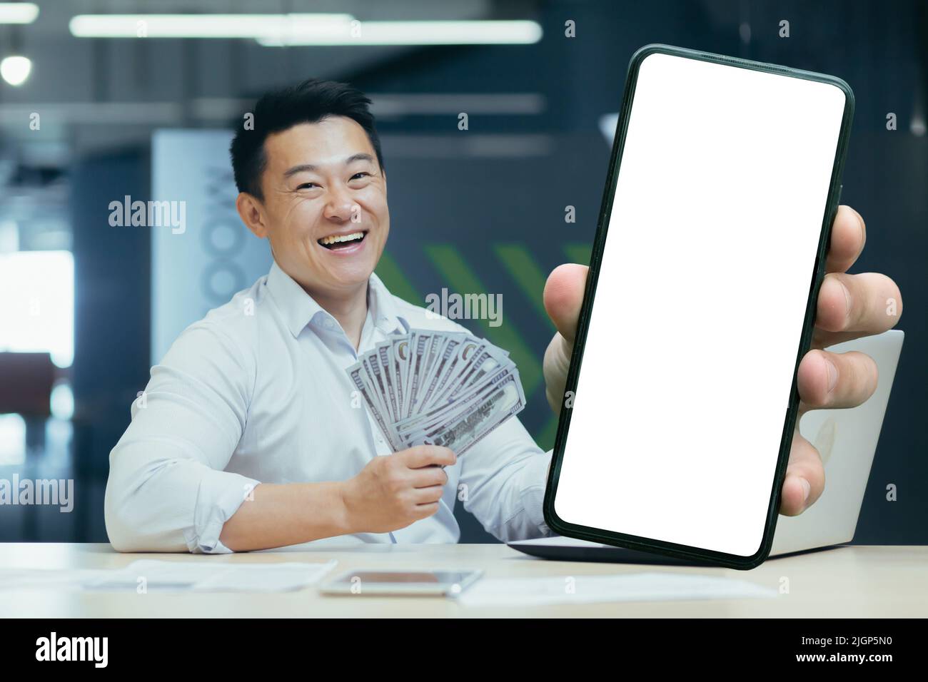 Asian felice uomo d'affari in ufficio, mostrando il telefono con schermo bianco, e tenendo denaro in mano, direttore boss felice con ricevuto prestito Foto Stock