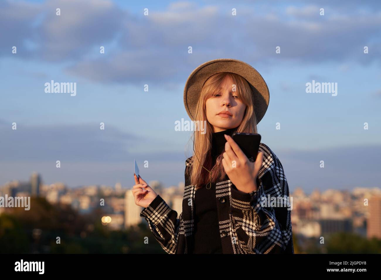 Attraente hipster ragazza turistica facendo shopping online con sfondo blu cielo nuvoloso e vista città. Giovane bionda caucasica in cappello con smartphone e carta di credito. Immagine di alta qualità Foto Stock