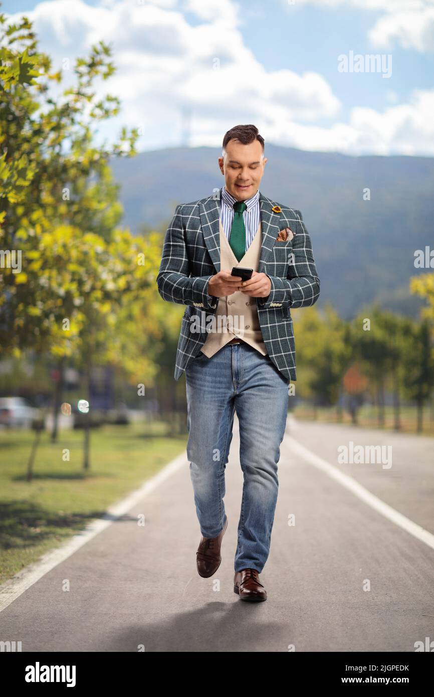Ritratto completo di un uomo in abito elegante e jeans che camminano su una corsia pedonale e utilizzando uno smartphone Foto Stock