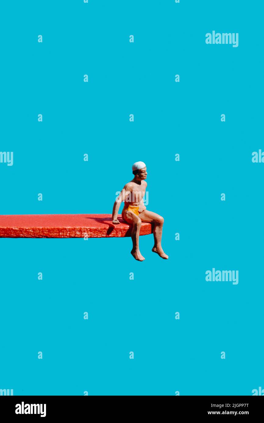 un uomo in miniatura, che indossa un costume da bagno giallo e un cappellino bianco, seduto su una tavola da immersione rossa, su sfondo blu Foto Stock
