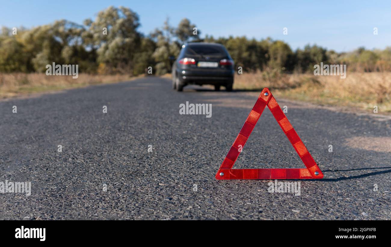 Triangolo segnaletico rosso sulla strada di fronte a un'auto rotta. Guasto della vettura in tempo di sole. Sicurezza del traffico stradale Foto Stock