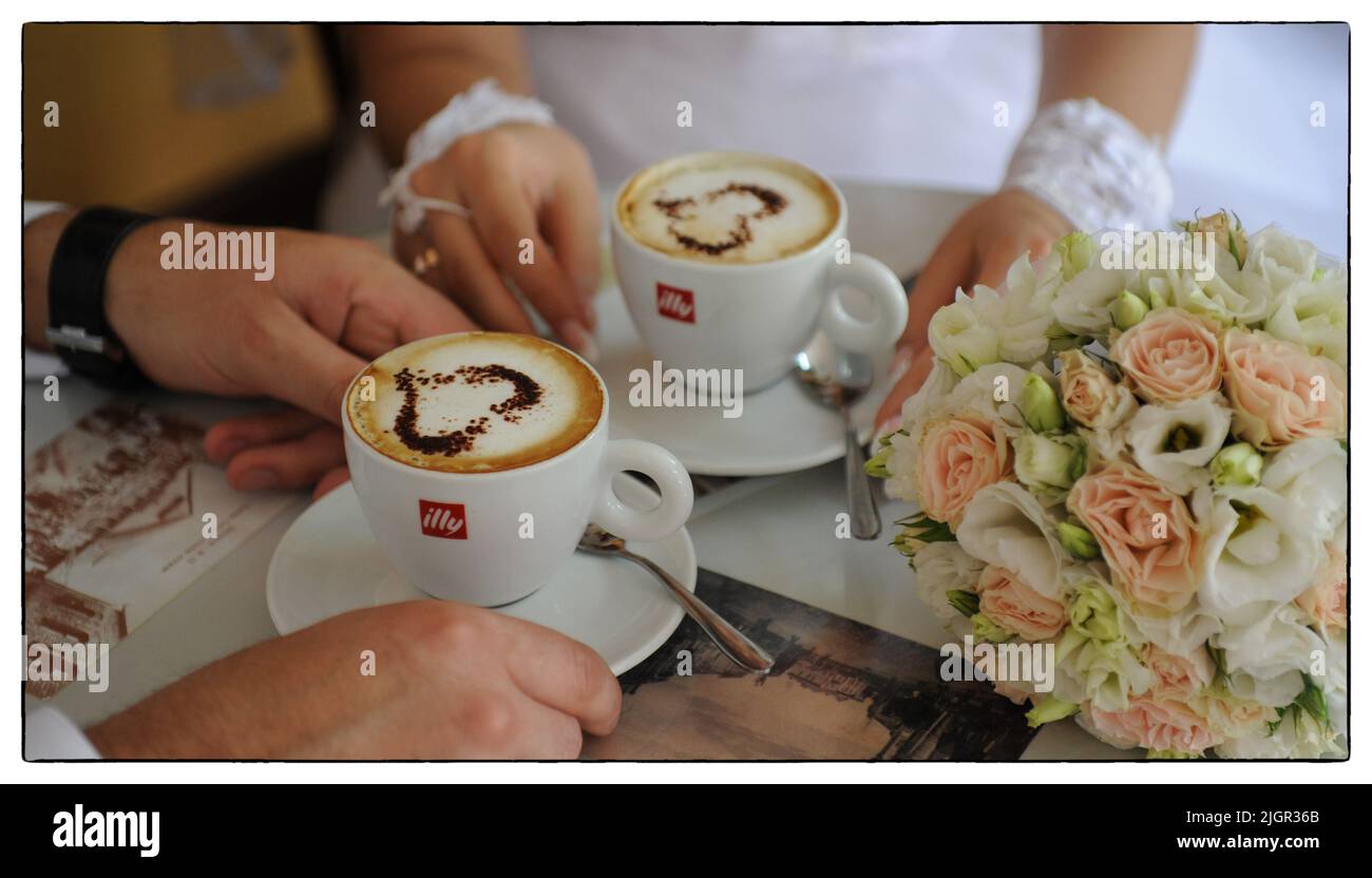 Le mani di nozze di un uomo e di una donna della sposa e sposo tengono nelle loro mani due tazze di caffè su cui c'è schiuma e un cuore disegnato su un tavolo con una tovaglia bianca. Foto di alta qualità Foto Stock