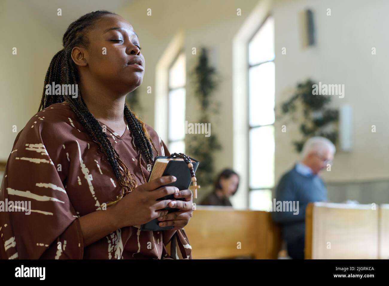 Giovane donna nera in abito casual mantenendo gli occhi chiusi durante la preghiera silenziosa in chiesa, tenendo la Sacra Bibbia e rosari perle Foto Stock