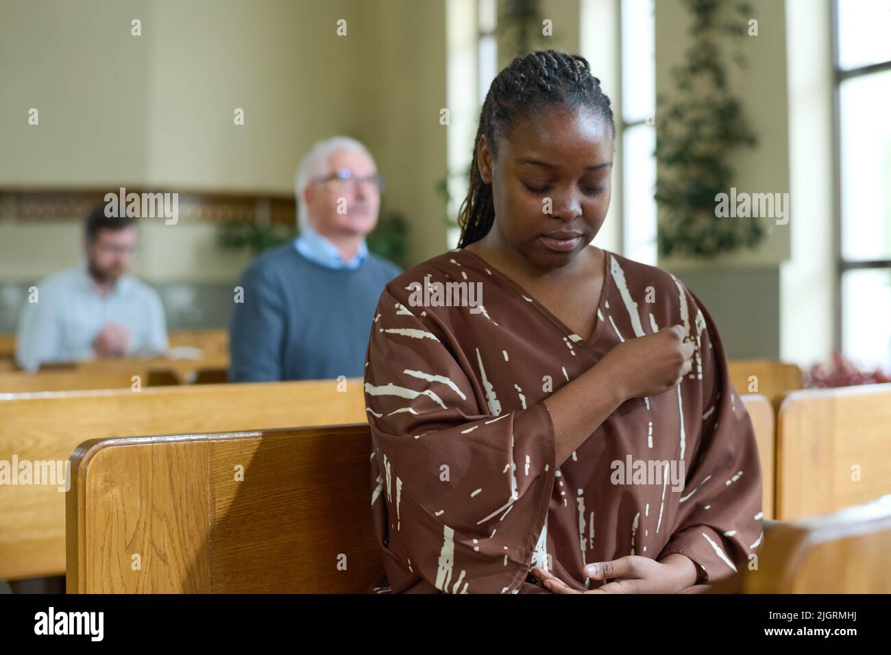 La giovane donna nera con gli occhi si è chiusa incrociandosi durante la preghiera silenziosa mentre si siede sul banco contro i parrocchiani in chiesa Foto Stock