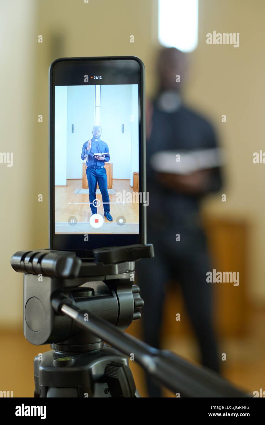 Schermo di smartphone con pastore che tiene aperta la Bibbia mentre si trova di fronte al pulpito di legno e predica durante il servizio della chiesa Foto Stock