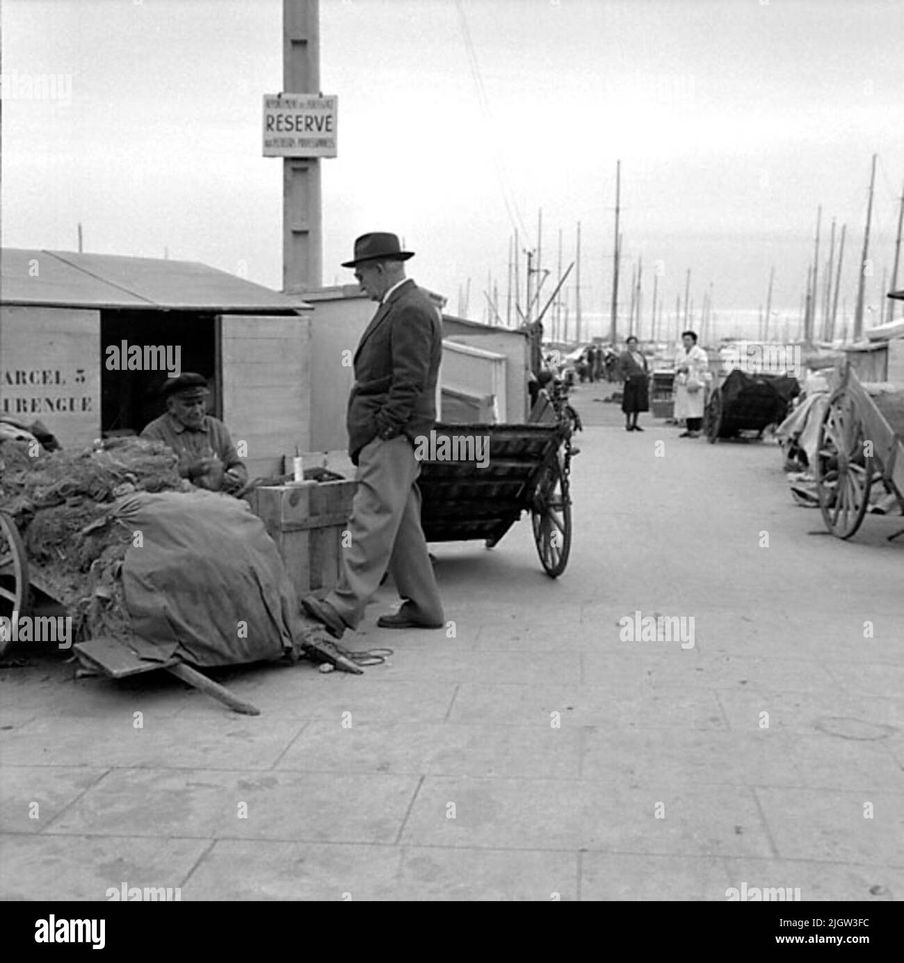 26. Francia. Il giornale fotografico è disponibile presso B.M.A. + Album fotografici. Acquisizione: Libri e materiale di archivio. Foto scattate 1959-11-05,12 Foto in serie. Secondo le note: Francia. Riviera, Cannes, 5/11 -59. Barche nel porto di pesca. Un uomo in giacca e cappello discute qualcosa con un uomo più anziano seduto, accanto ad un carro con reti da pesca ecc più lontano, due donne attendono. Dietro di loro c'è un certo numero di alberi di barca. Foto Stock