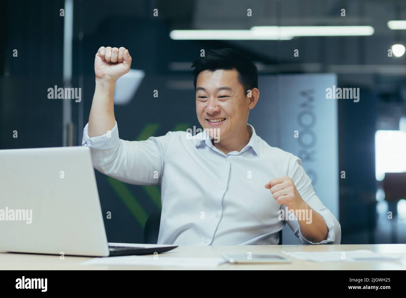 Allegro e riuscito uomo d'affari asiatico ballare in ufficio celebrare il trionfo e la vittoria seduta alla scrivania di lavoro con il portatile Foto Stock