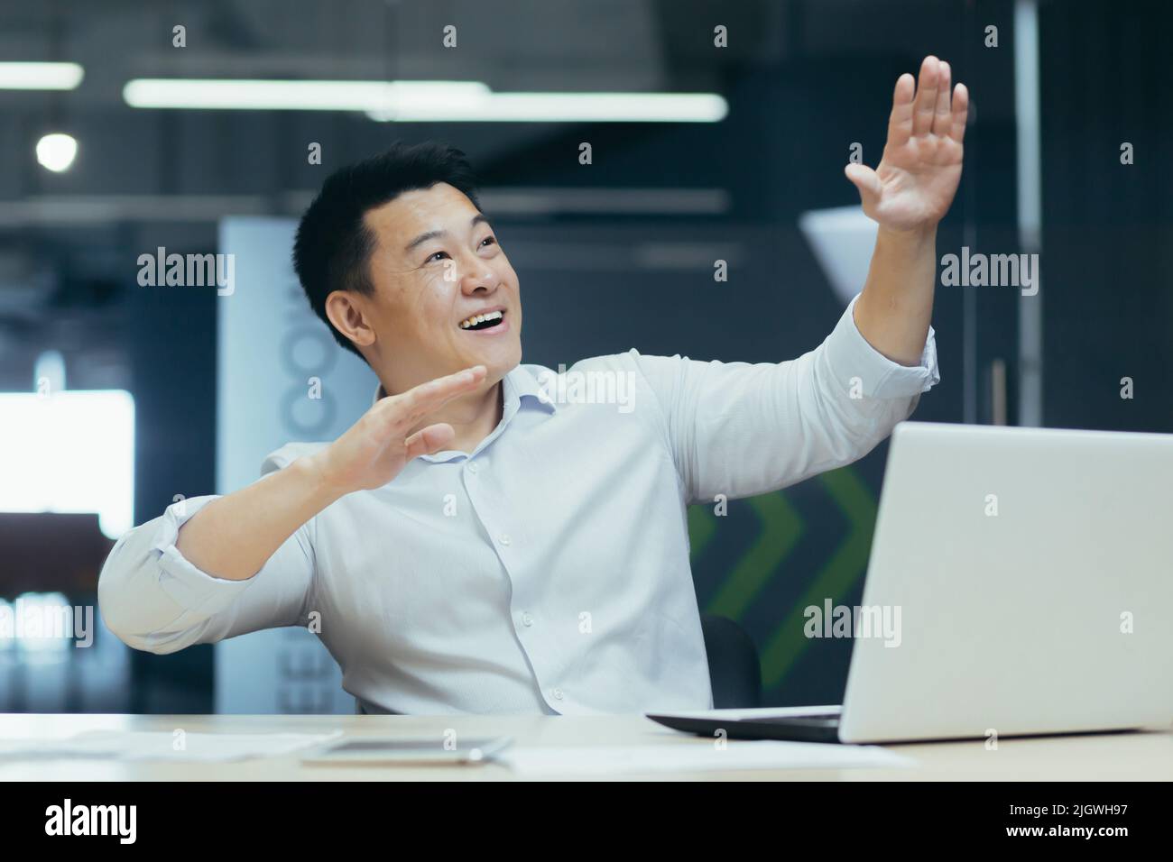 Allegro e riuscito uomo d'affari asiatico ballare in ufficio celebrare il trionfo e la vittoria seduta alla scrivania di lavoro con il portatile Foto Stock