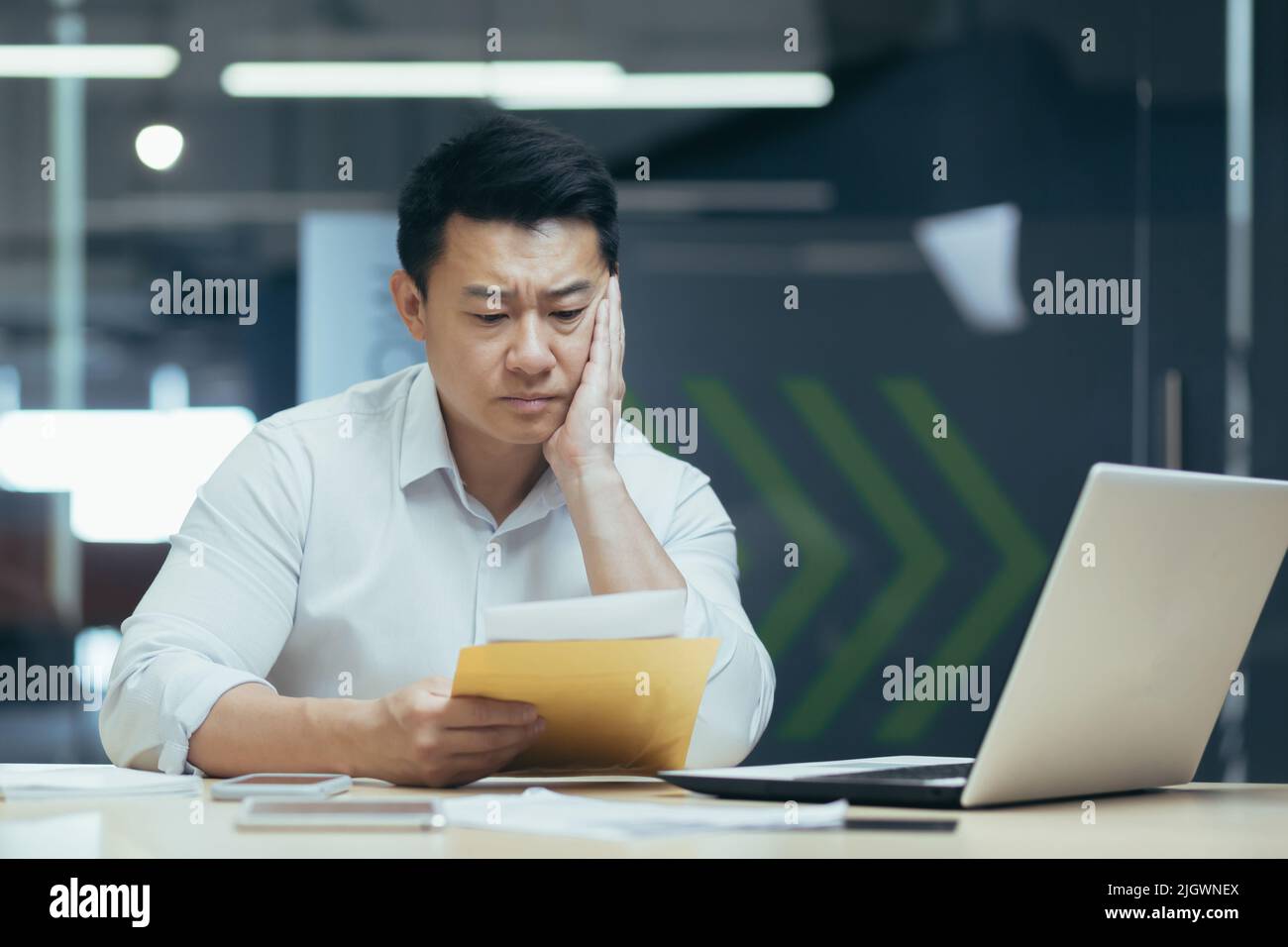 Triste sopra pensare l'uomo d'affari asiatico che legge la lettera difettosa di notizie dalla banca, capo maschio in camicia che funziona in ufficio Foto Stock