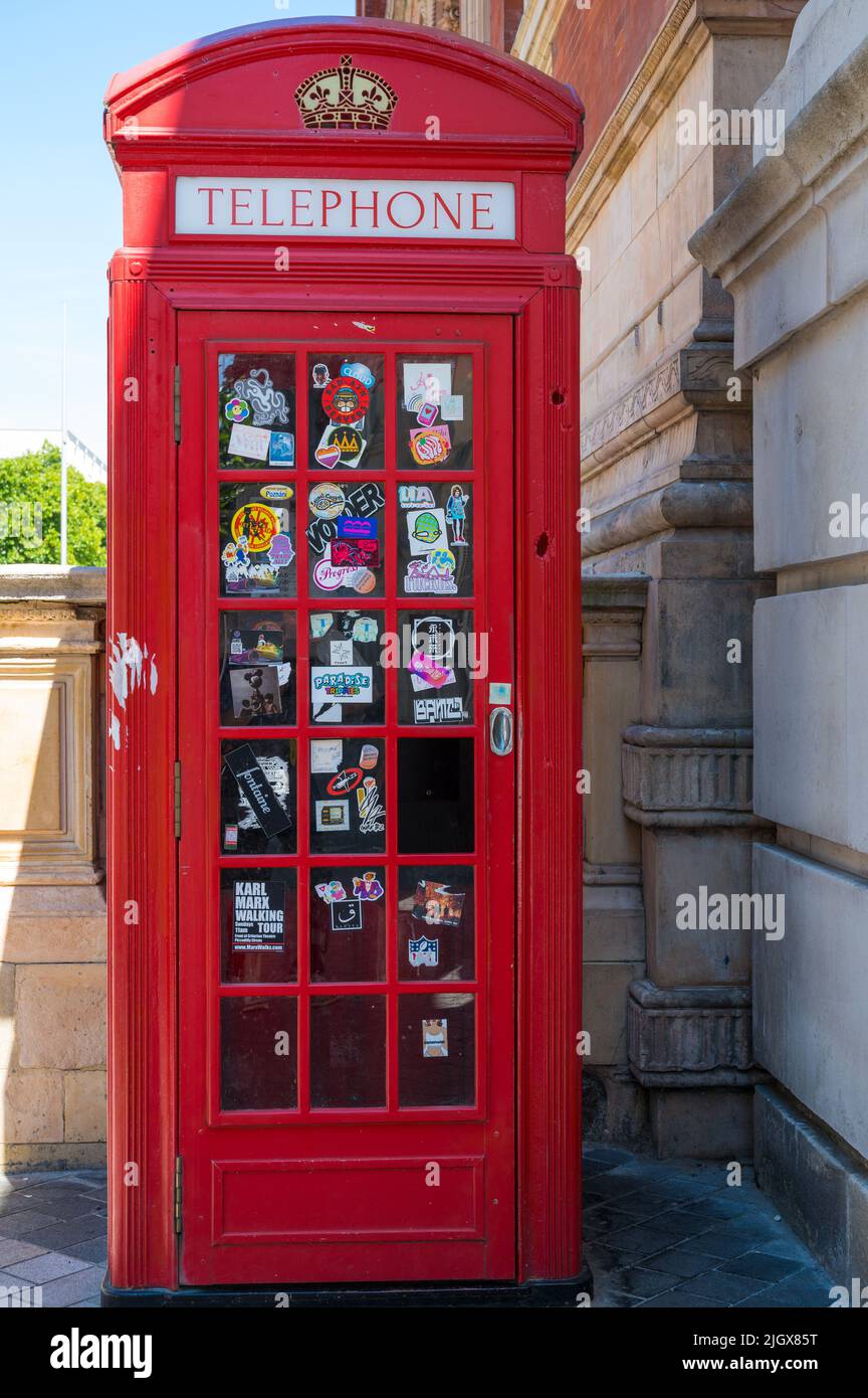 Telefono tradizionale rosso con vari adesivi pubblicitari sui vetri delle porte. Exhibition Road, South Kensington, Londra, Inghilterra, Regno Unito Foto Stock
