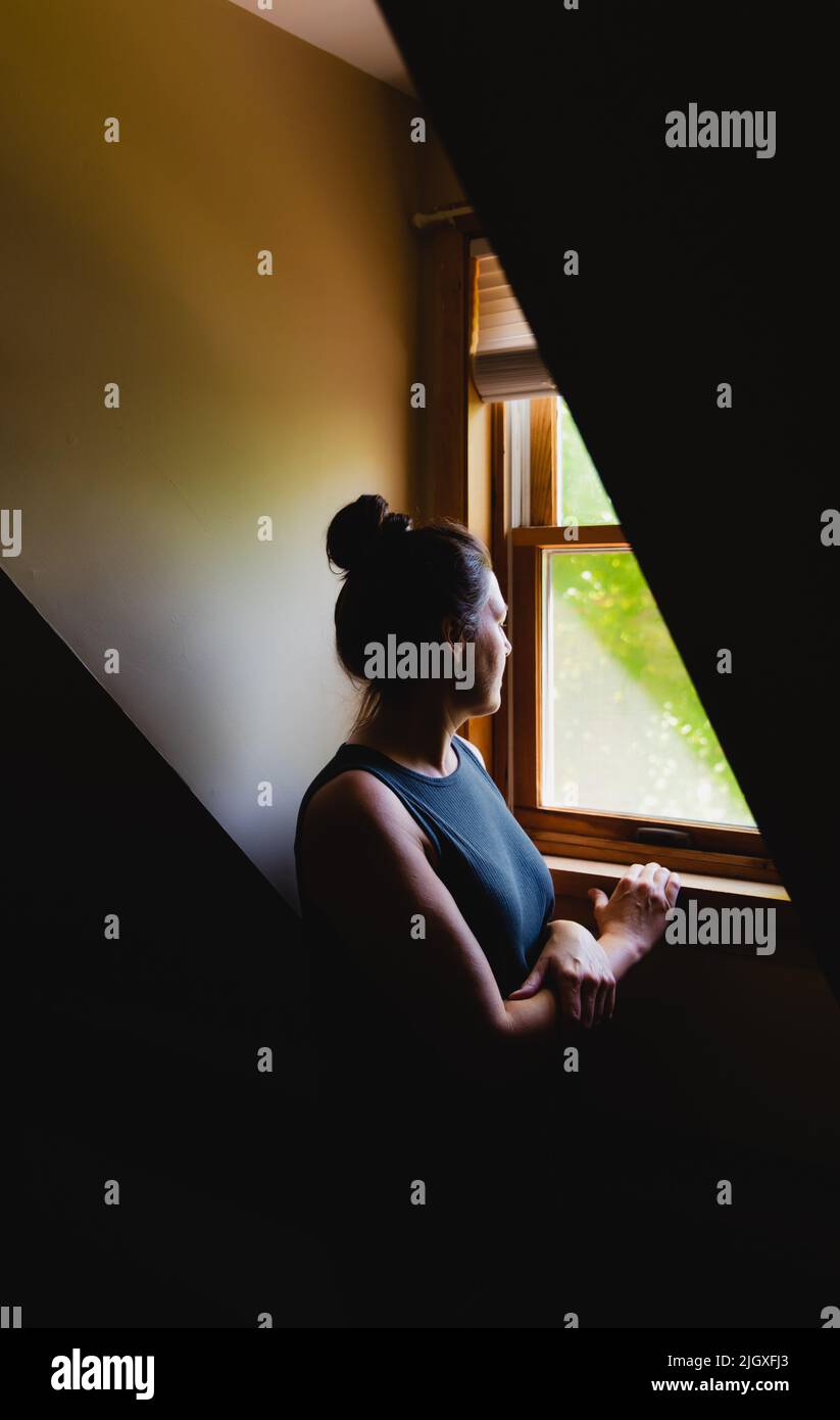Donna che guarda da una finestra dormitorio in una stanza buia di giorno. Foto Stock