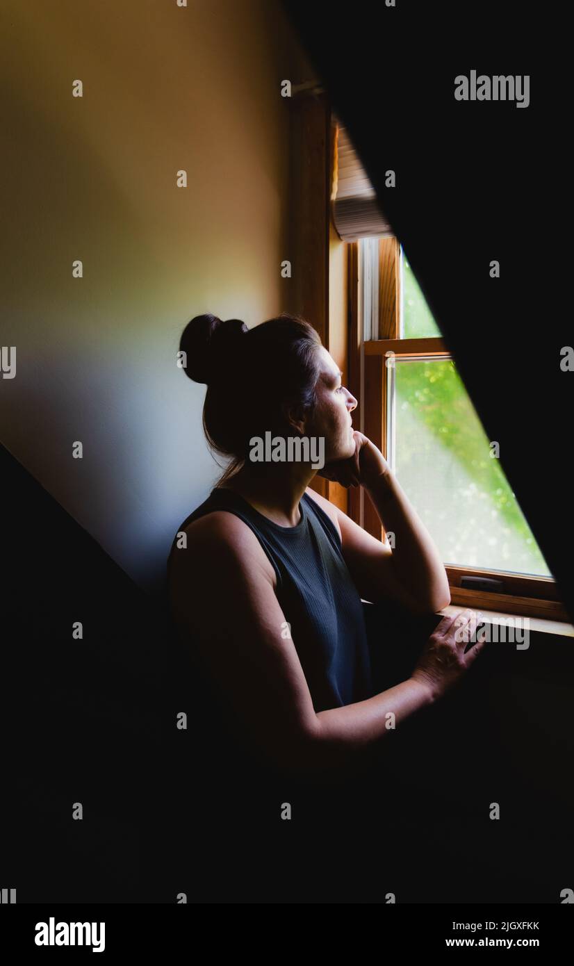Donna che guarda da una finestra in una stanza buia di giorno. Foto Stock